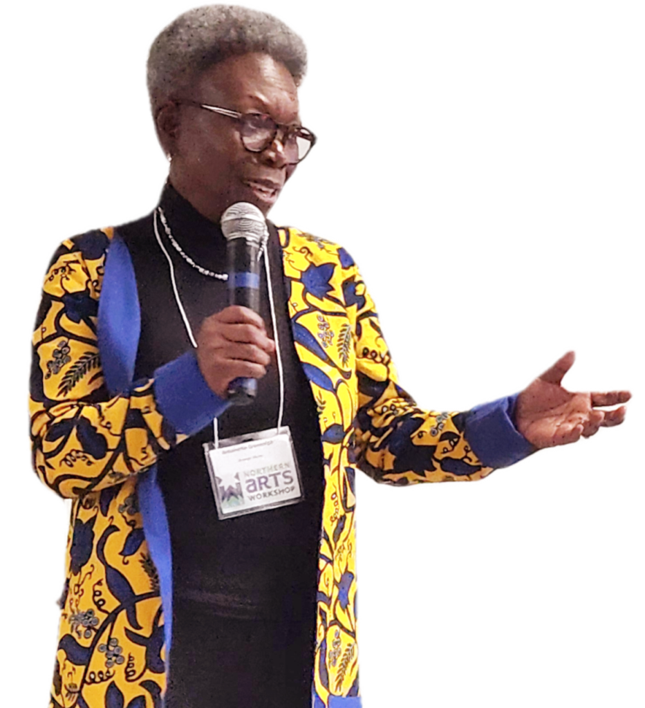 Antoinette GreenOliph, keynote speaker at the Northern Arts and Food Workshop in The Pas, MB on March 325, 2024. Black woman with microphone speaking, upper body and head wearing a yellow and blue long jacket and black top.