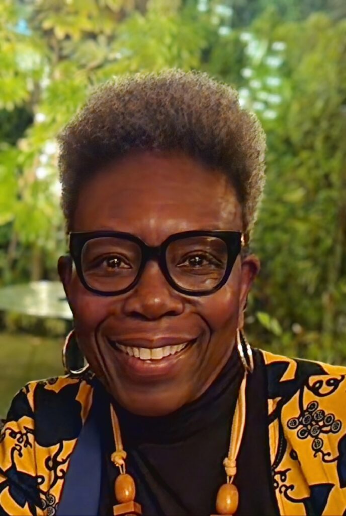 Antoinette GreenOliph, keynote speaker, entrepreneur, mentor and storyteller. Black woman wearing glasses, a yellow and blue jacket and black top, smiling at the camera with a green treed background.